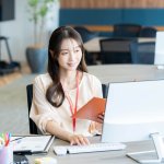 ファイルを片手にデスクでPC作業をするOL、スーツ、女性