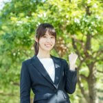 木漏れ日の中でガッツポーズをするスーツ姿のポニーテールの女性、OL