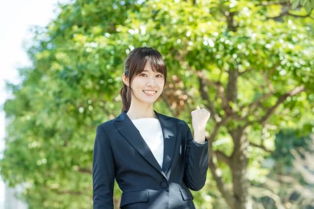 木漏れ日の中でガッツポーズをするスーツ姿のポニーテールの女性、OL